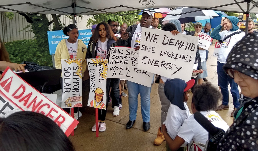 Several people gather under tents and umbrellas holding signs that say, "We demand safe affordable energy," "Green Economy," "Support solar," and more