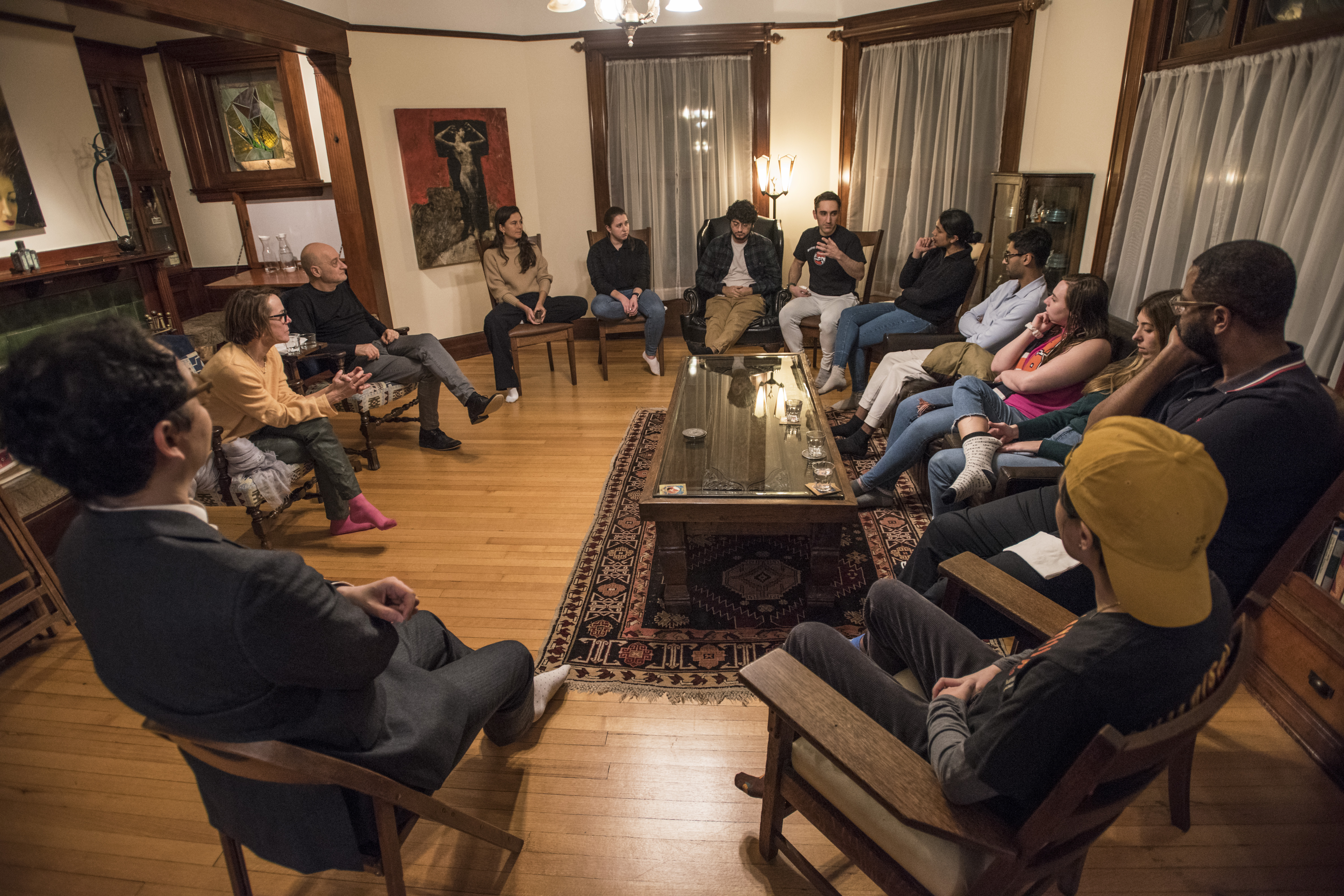 The class sitting on chairs in a circle, listening to a speaker. Location seems to be a living room.