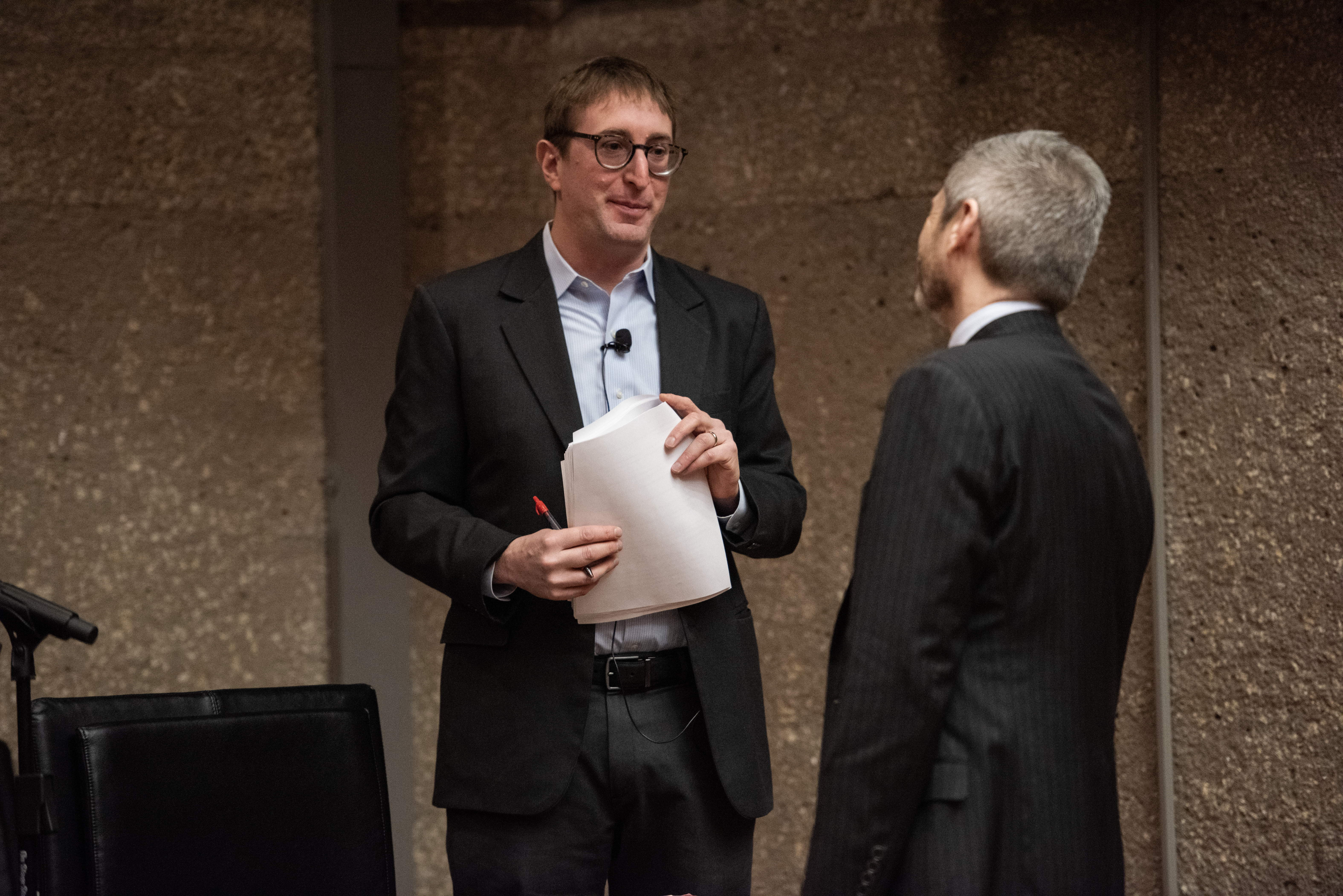 A man wearing a suit and holding papers stands in front of another man as they engage in a brief exchange.