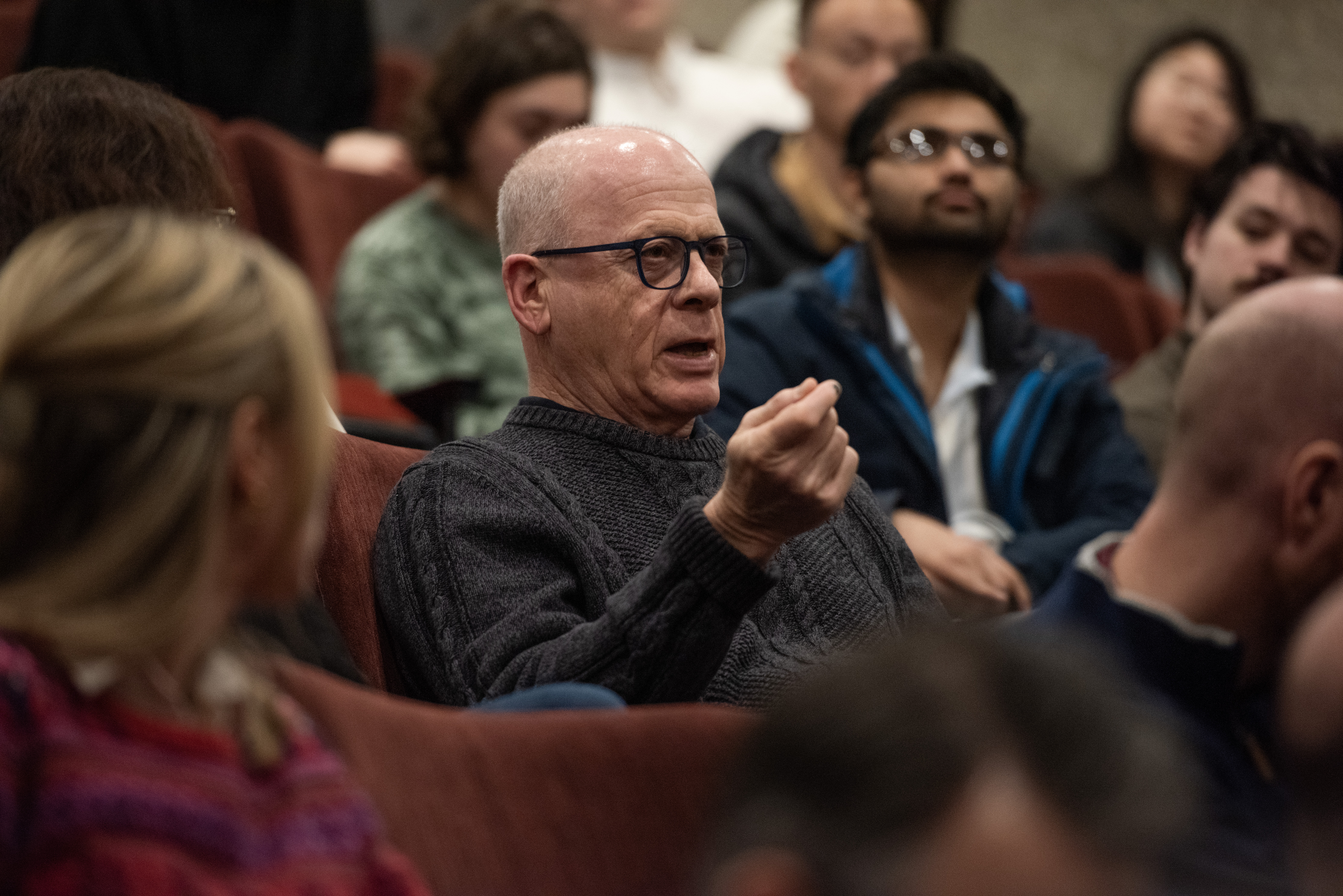 A man sits in a row of seats filled with other people (students) and appears to be asking a question to Jacob Goldin who is not pictured.