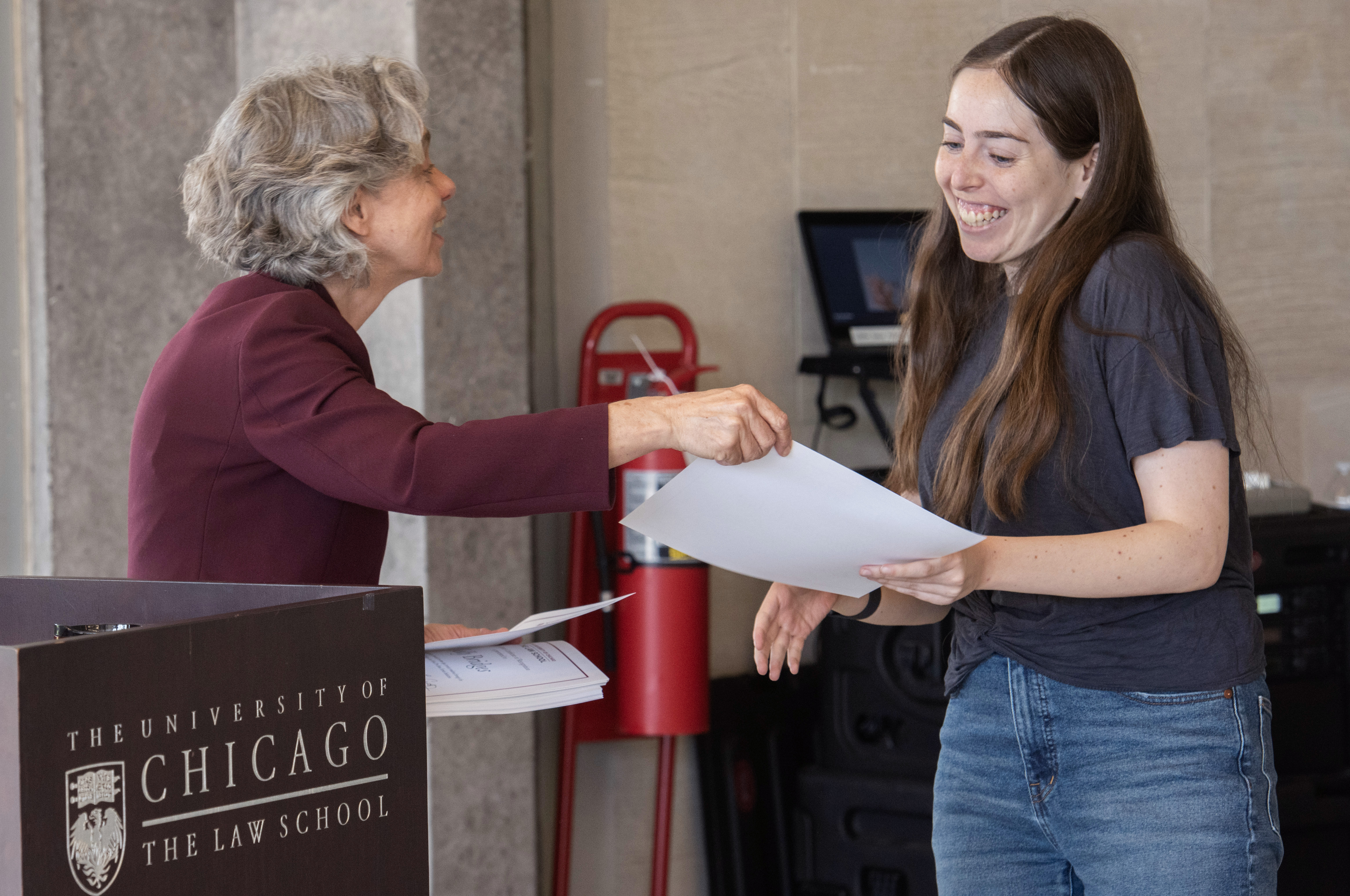 A woman standing near a lectern with a microphone hands a paper to another woman who receives it smiling.