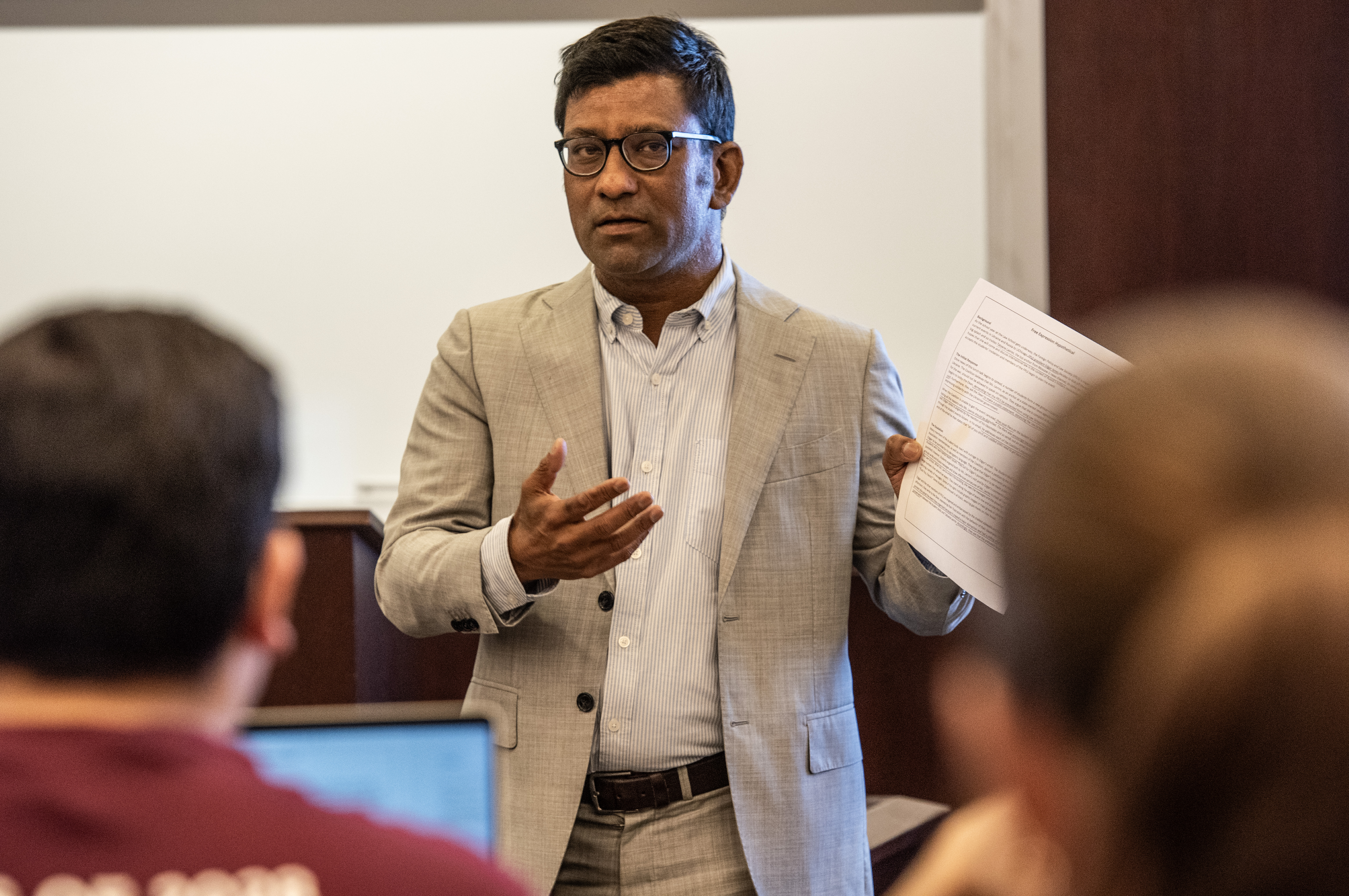 A male professor wearing a suit gestures as he speaks to a class of students.