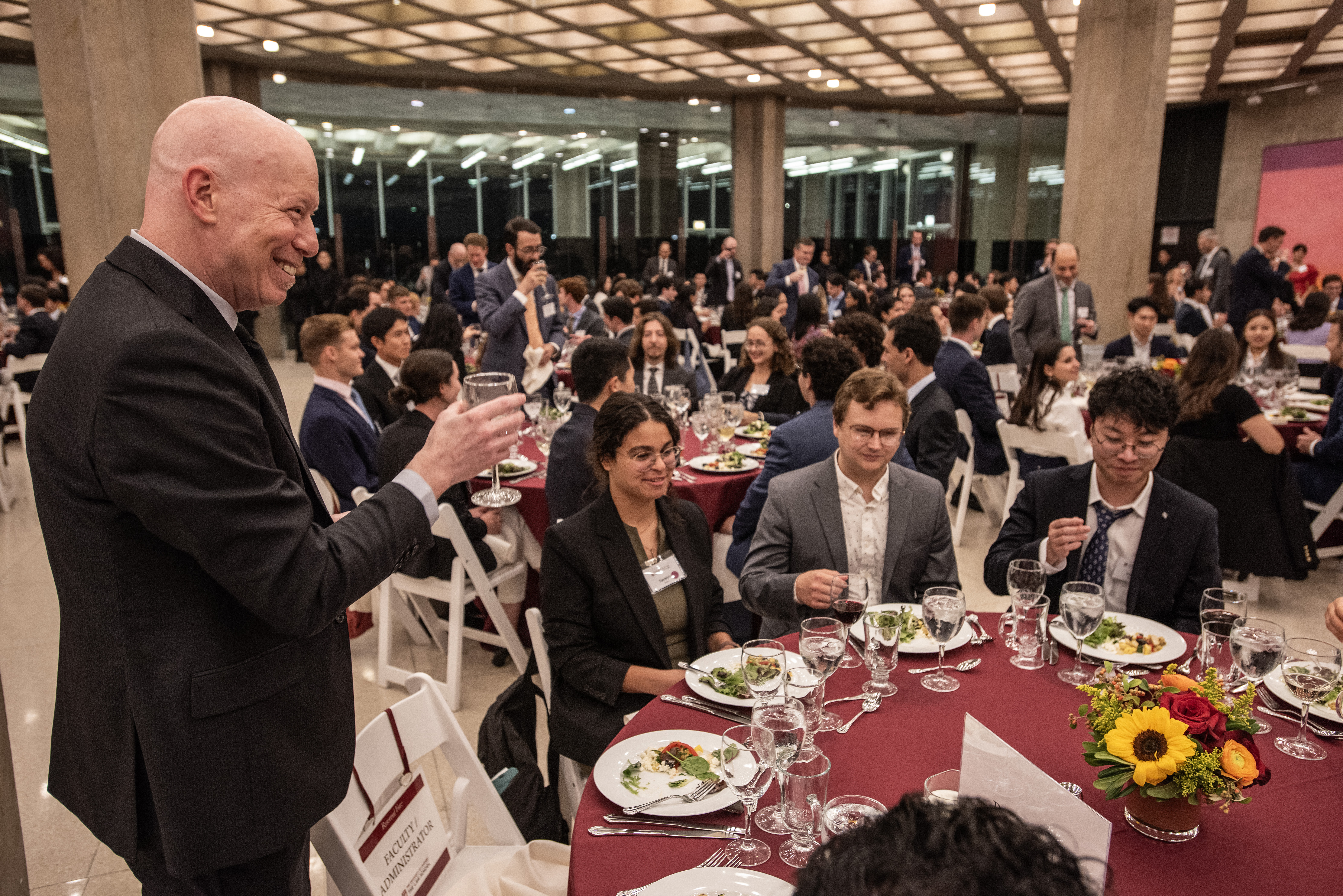 A male professor dressed in a suit stands next to one of many round dinner tables in a large room raising his glass to cheers with students who are seated at the dinner tables.