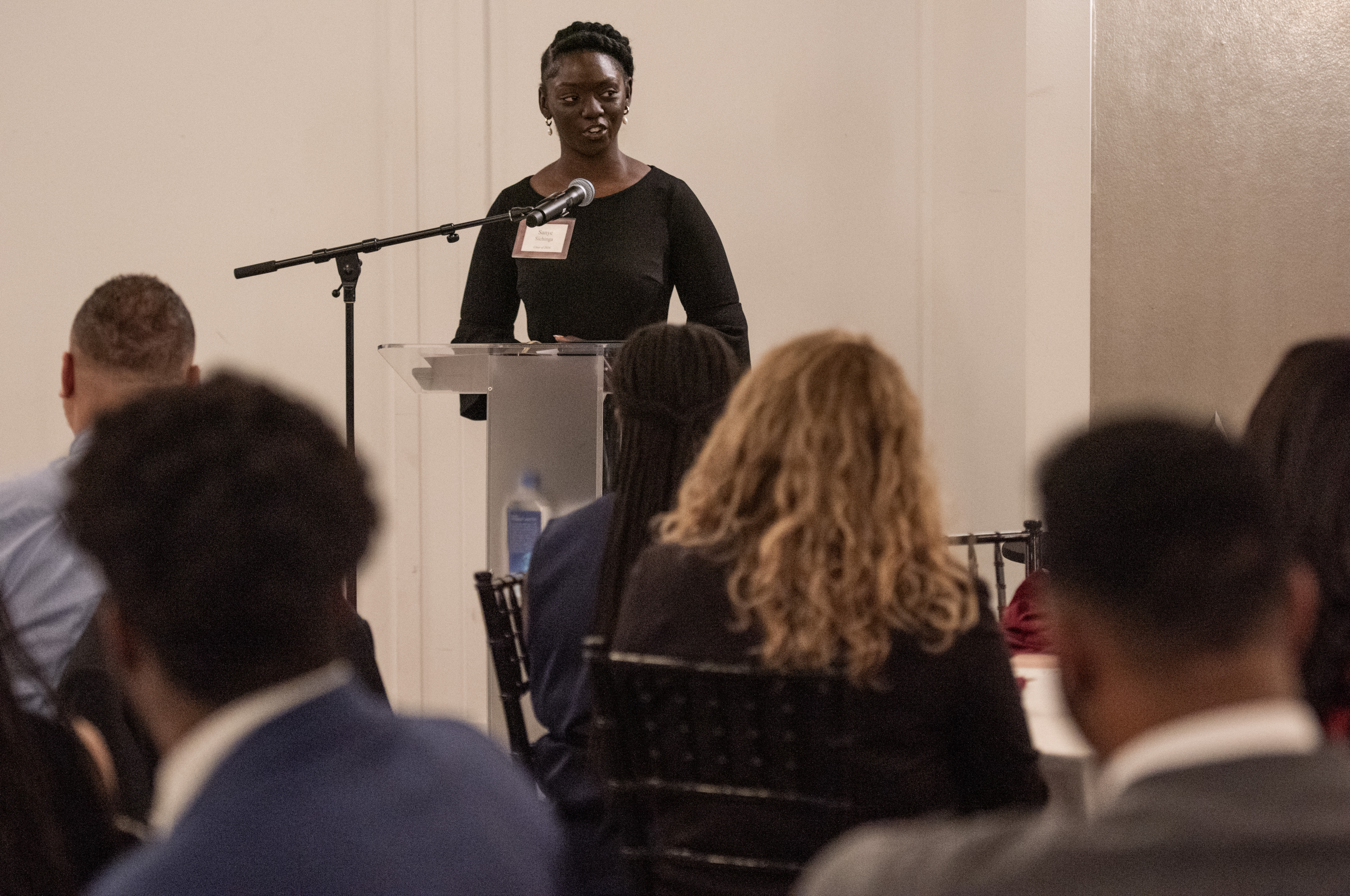 A young Black woman dressed elegantly speaks at a podium in front of a crowd.