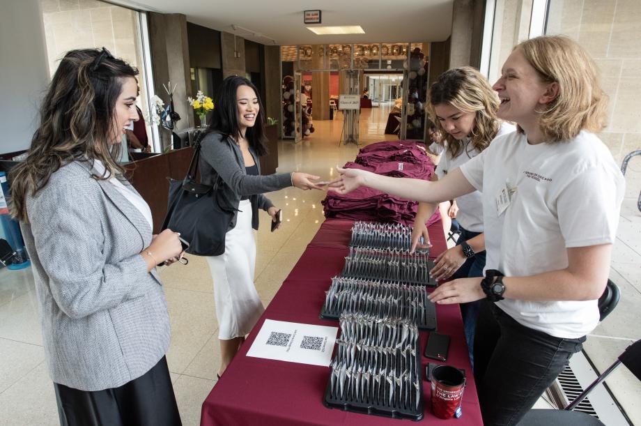 Two students in white shirts stand on one side of a table handing out nametags. Two new students on the other side of the table collect their nametags.