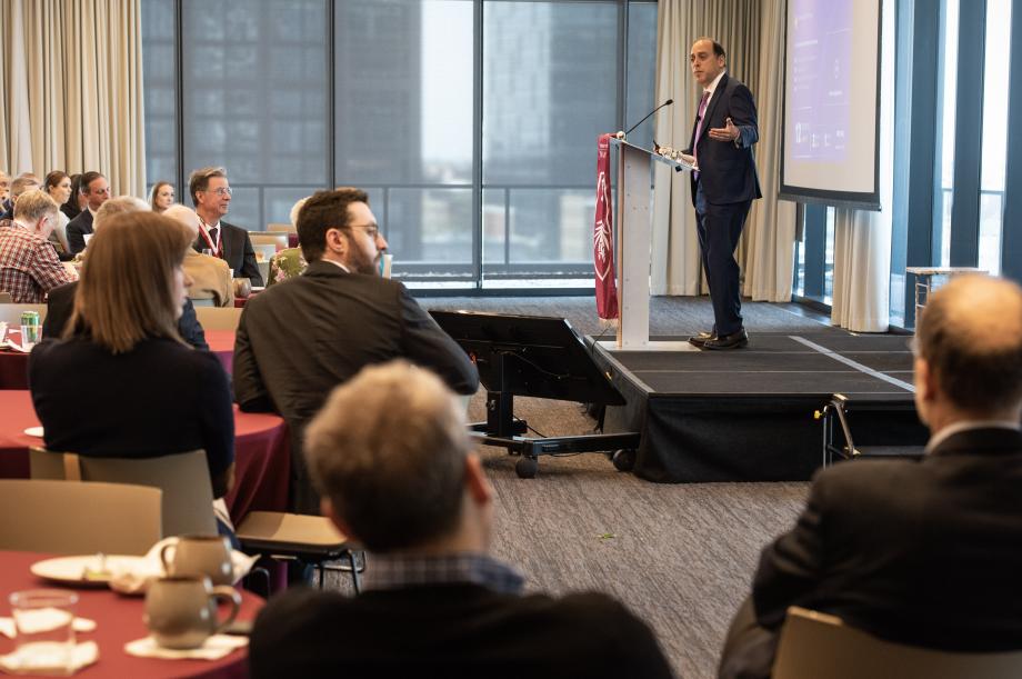 Professor Strahilevitz stands behind a podium on a stage with people at round tables looking up at him.