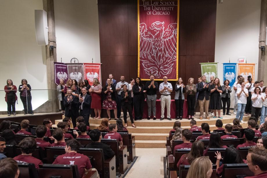 Approximately two dozen staff members and orientation leaders stand on a stage in the auditorium. New students wearing matching maroon shirts are seated in the foreground.