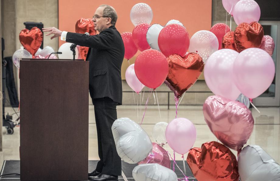 Douglas Baird, in a tuxedo, stands at a podium with red, pink and white balloons behind him.