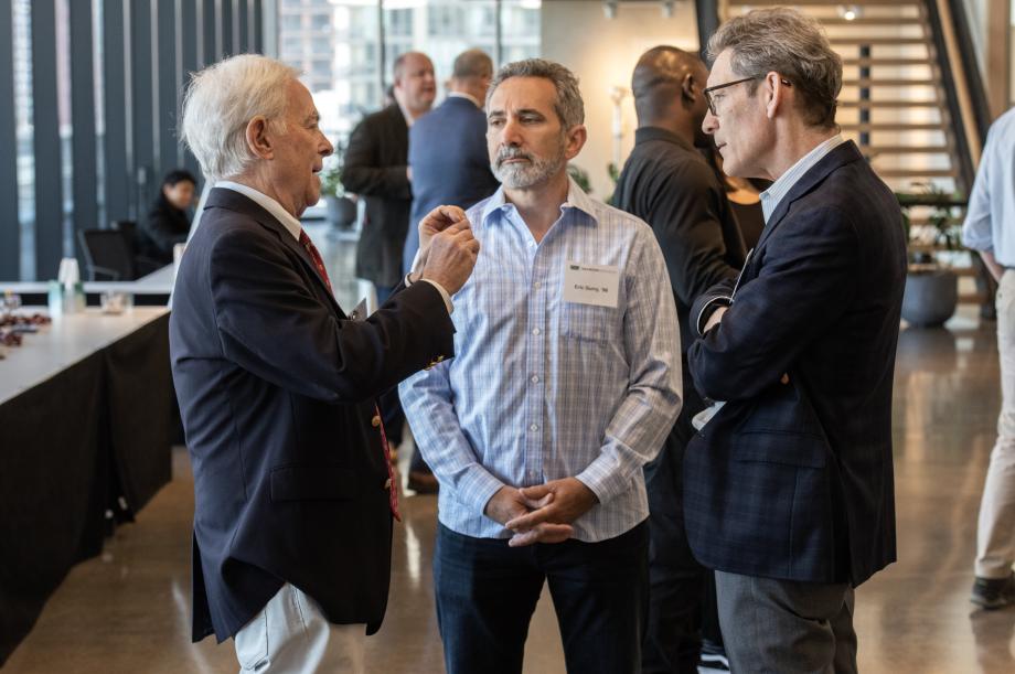 A man gesticulates as two other men look on. Additional people are in the background.