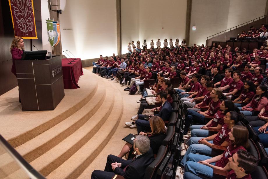 Brandi Welch, wearing a maroon suit, stands at the podium with a sea of students seated in front of her.