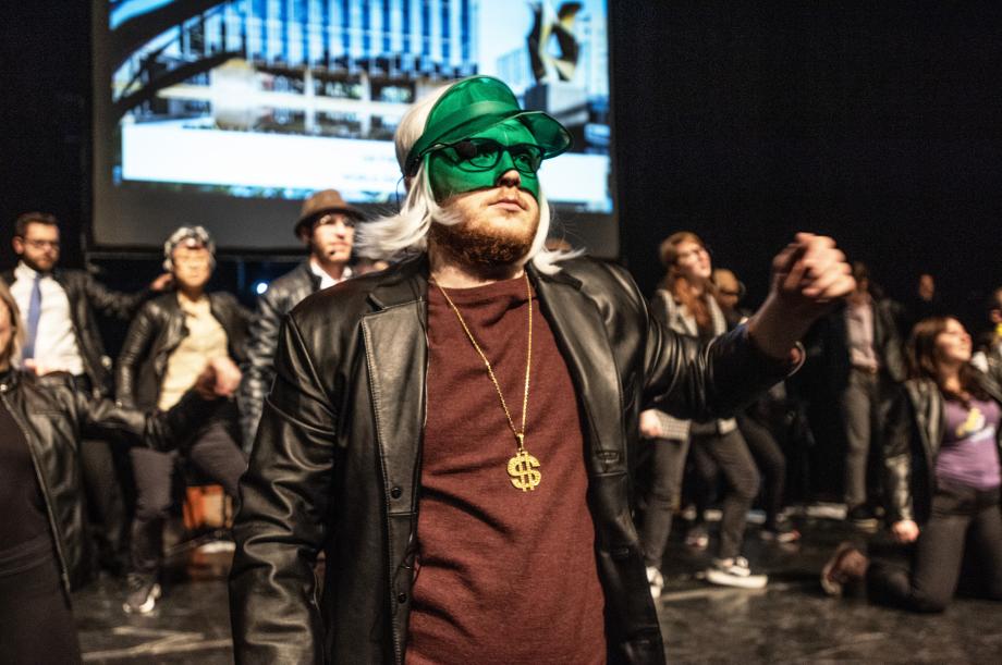 A group of people wearing leather jackets, with particular focus on a person who also wears a partial green mask and a gold necklace. 