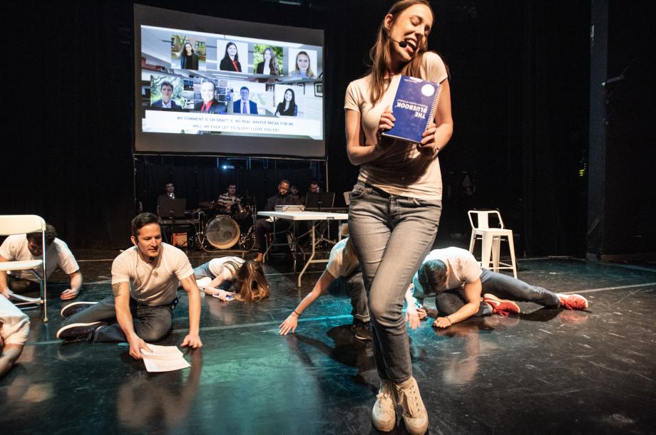 People fallen on the floor while the journal staffer sings and holds a blue book in front of them.