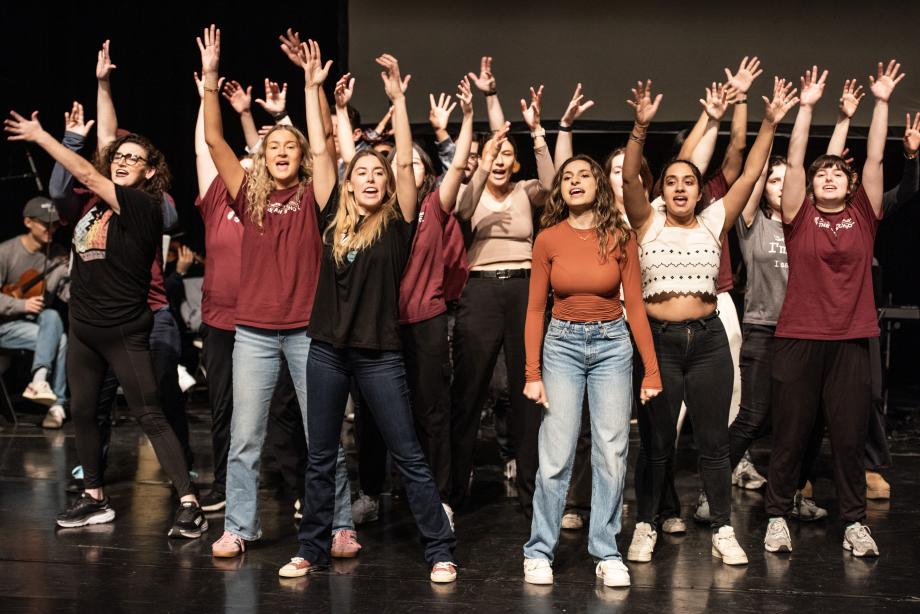 A group of people standing on a stage visibly singing with their hands outstretched and raised above their heads.