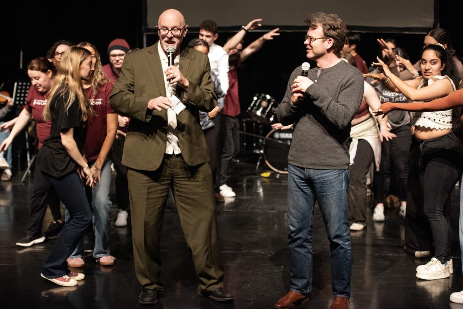 Professor Ginsburg and Professor Birdthistle are standing center stage holding microphones while students are moving and waving their arms behind them.