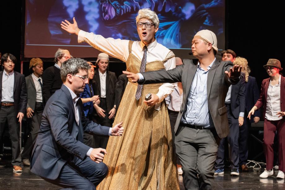 A man wearing a gold dress and a grey wig is singing center stage; on either side of him is a person wearing a grey suit and dancing; behind the trio are a group of people standing and watching.