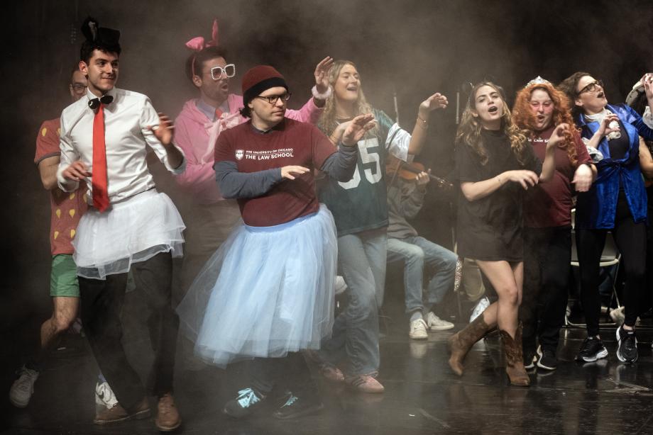 A group of people wearing costumes such as skirts and oversized glasses are dancing in a thin cloud of smoke.