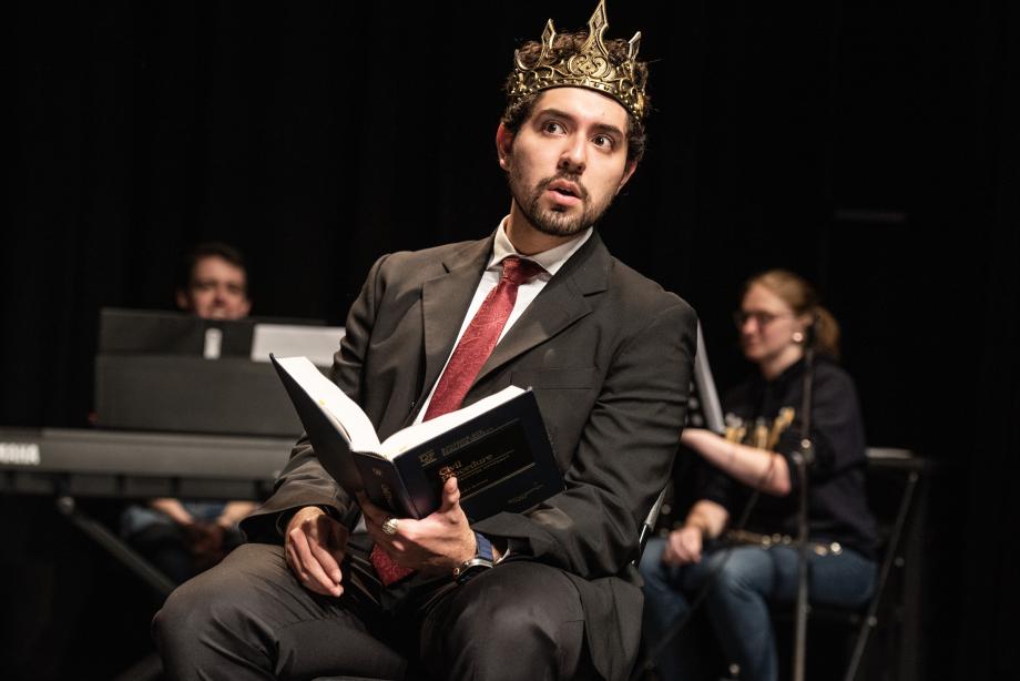 A man wearing a gold crown on his dead, a dark suit, and a red tie is sitting on a chair, holding an open textbook in one hand, while his eyes are looking cautiously to the side.