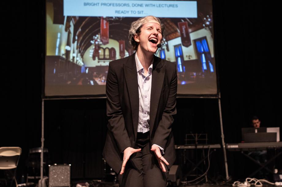 A person wearing a grey wig and a black blazer is belting out a song on stage with their eyes closed.