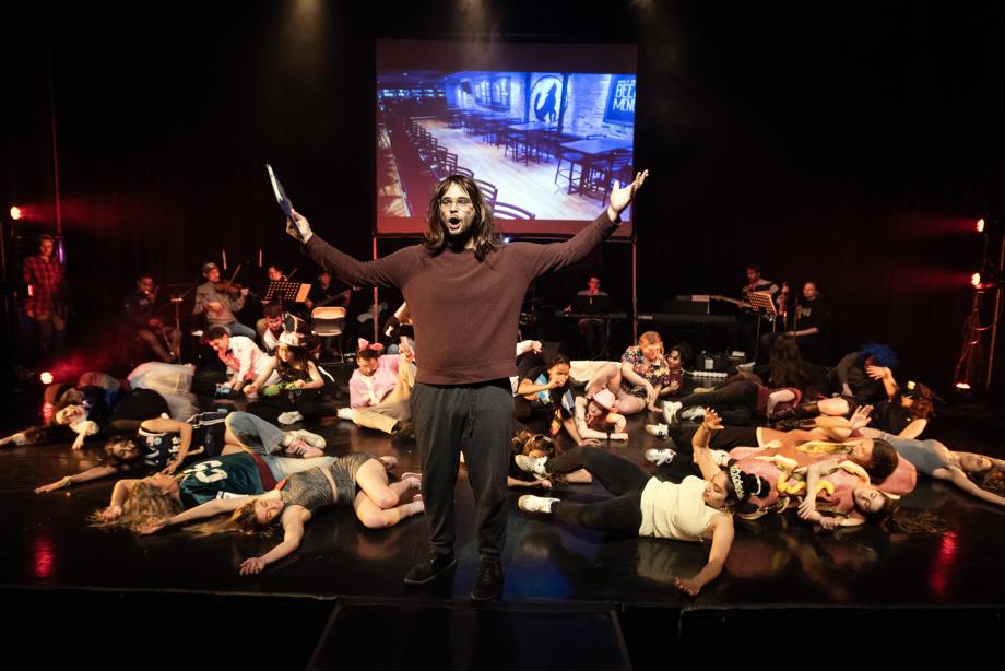 A man wearing a shoulder-length brown wig is standing center stage singing with arms raised; behind him are people sprawled out on the floor.