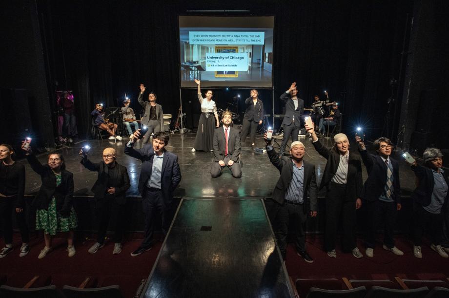 A wide shot of the entire stage that shows a group of people lined up and waving; in the middle of the stage is a man kneeling on the floor with a gold crown on his head.