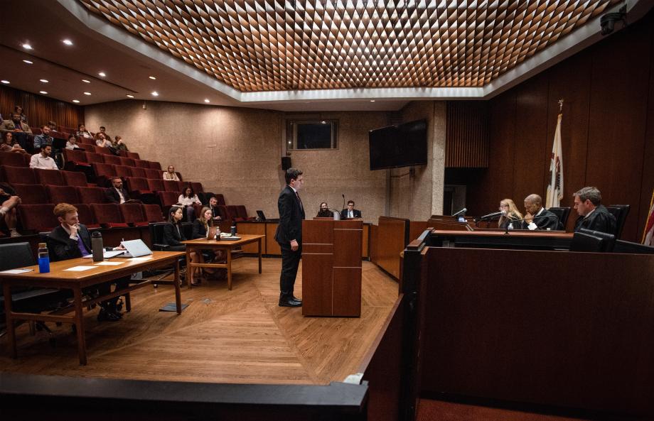 A student stands at the podium in the Weymouth Kirkland Courtroom before a panel of three judges seated at the bench. Two desks with students are behind the podium, and the audience looks on.