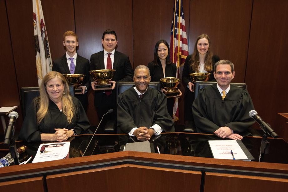 Four students holding their trophies pose behind the three judges at the bench.