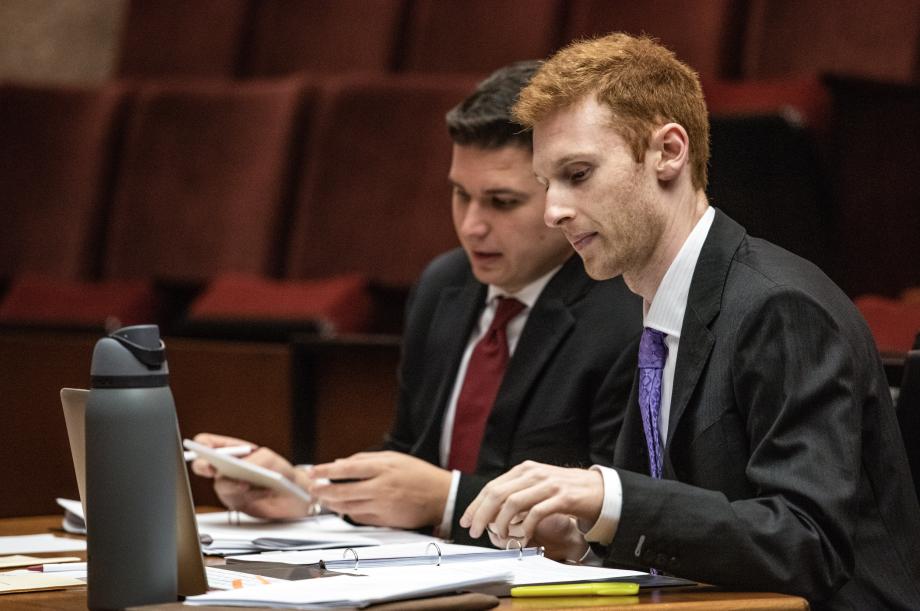 Two students in suits sit at desk with binders, papers, and a laptop strewn in front of them.