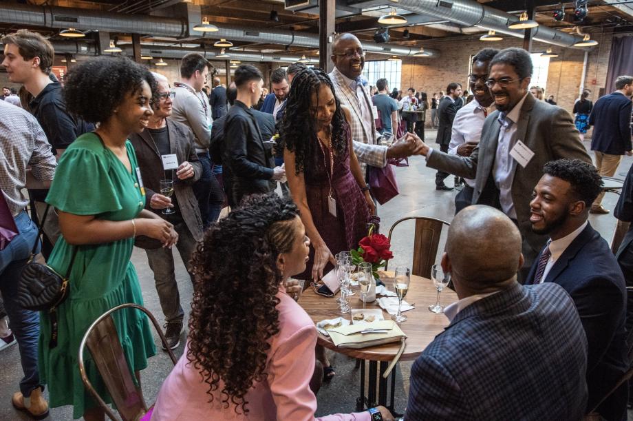 Three people are seated at a small top table while five people stand around them in a former warehouse space.