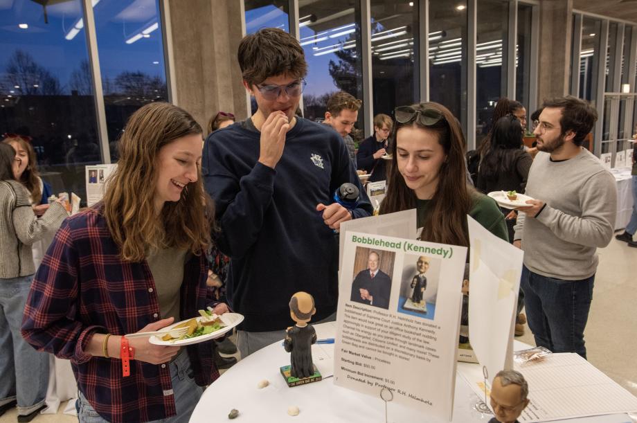 Three students standing behind a round table facing the camera and looking at a bobblehead. A sign on top of the table says "Bobblehead (Kennedy)".
