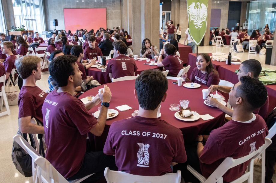 New students in matching maroon shirts sit around large circular tables.