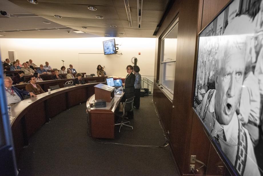 Two professors stand at the front of a classroom with alumni seated in tiered semi-circle rows. A clip from a black and white film appears on the screen in the foreground.