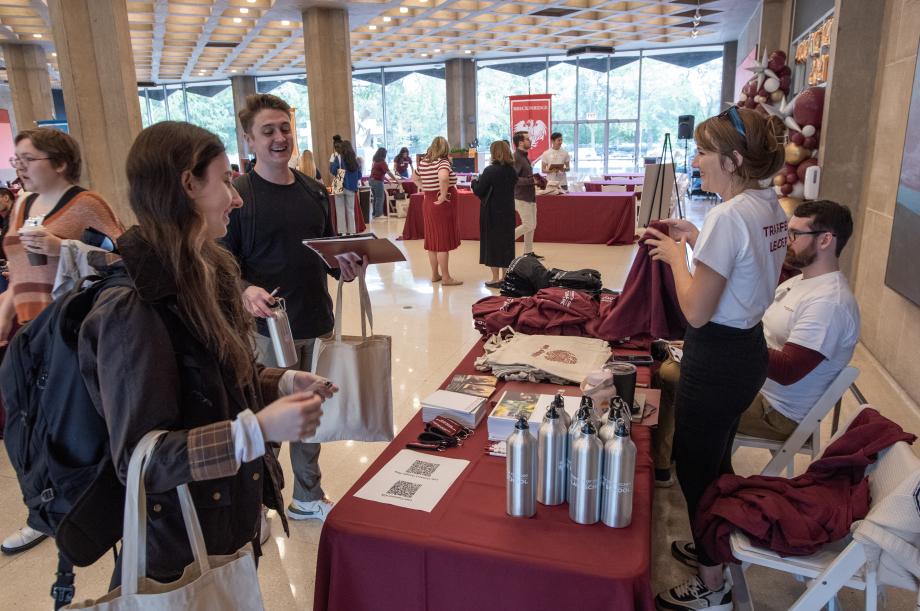 Two orientation leaders in white shirts stand and sit behind a table with swag items as two new students with water bottles and tote bags chat.