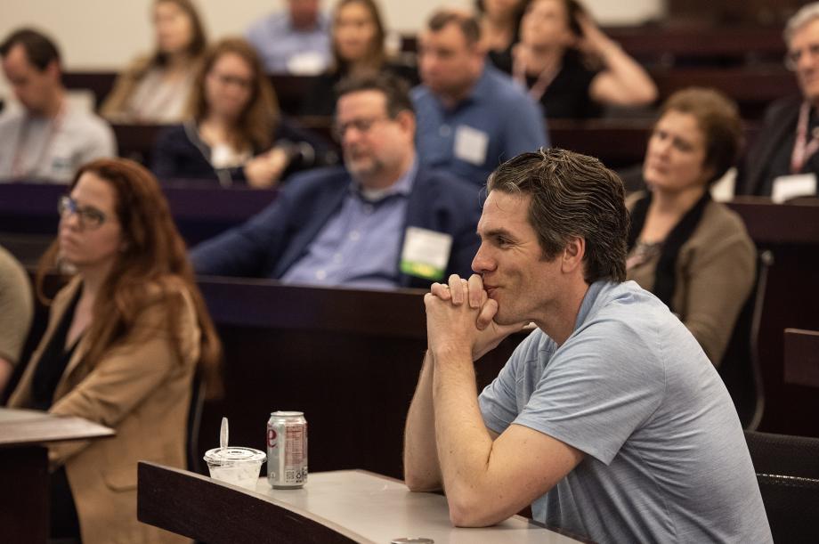 A man in a t-shirt leans against his arms on the desk in front of him. Other alumni, slightly out of focus, look to the front of the classroom.