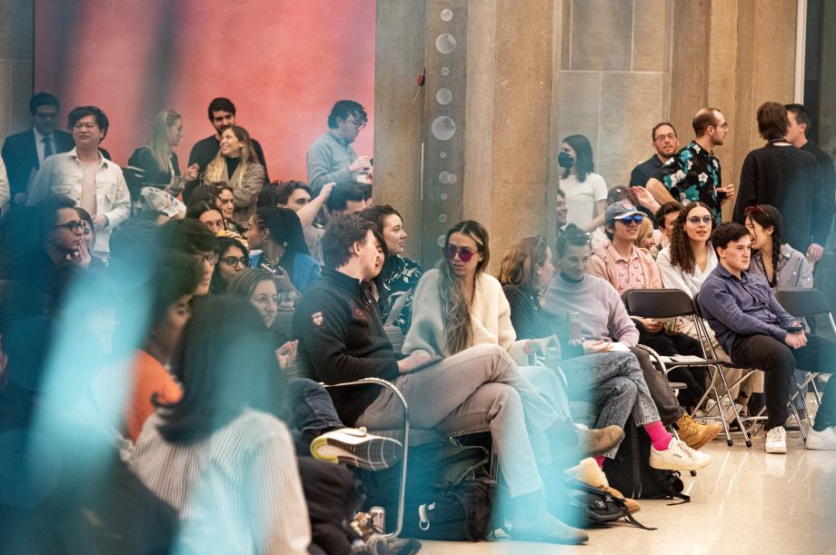 Students sitting in a large, crowded space surrounded by faint blue lights.