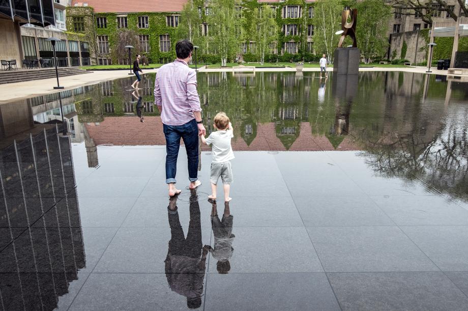 A barefoot man, holding on to a barefoot child venture into 1 inch deep water reflecting the sky and nearby buildings.