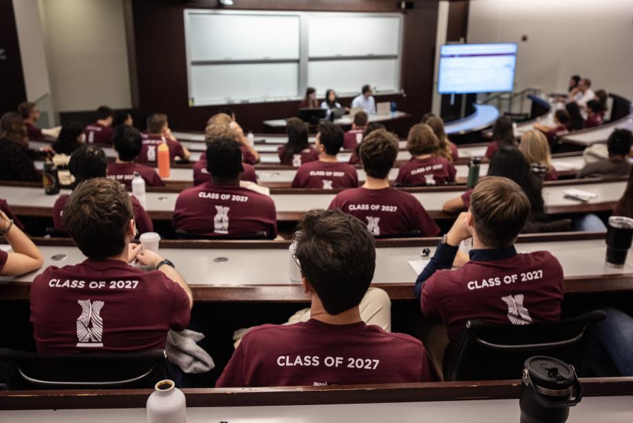 Students with maroon shirts reading, “Class of 2027” on the back, sit in a classroom looking at a three-person panel at the front of the room.