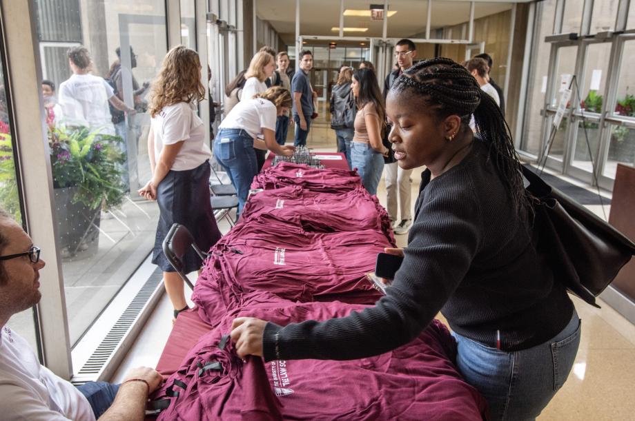A new student in a black sweater picks out a maroon shirt from a table while other new students line up to receive their nametags.