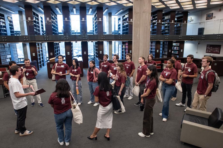 Approximately 20 new students gather in a circle around an orientation leader in a white shirt. Library stacks appear behind the group.