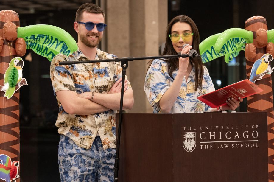 Two individuals at the podium wearing tropical outfits and sunglasses. The speaker is holding a red clipboard.