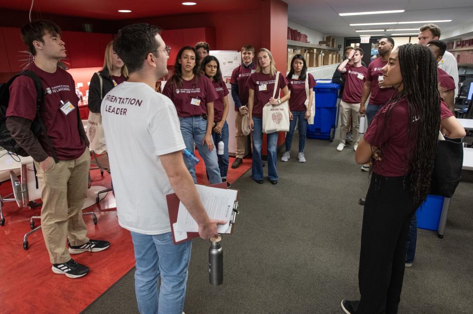 Approximately 15 new students gather in a circle around an orientation leader in a white shirt. They are in an office and kitchenette space.