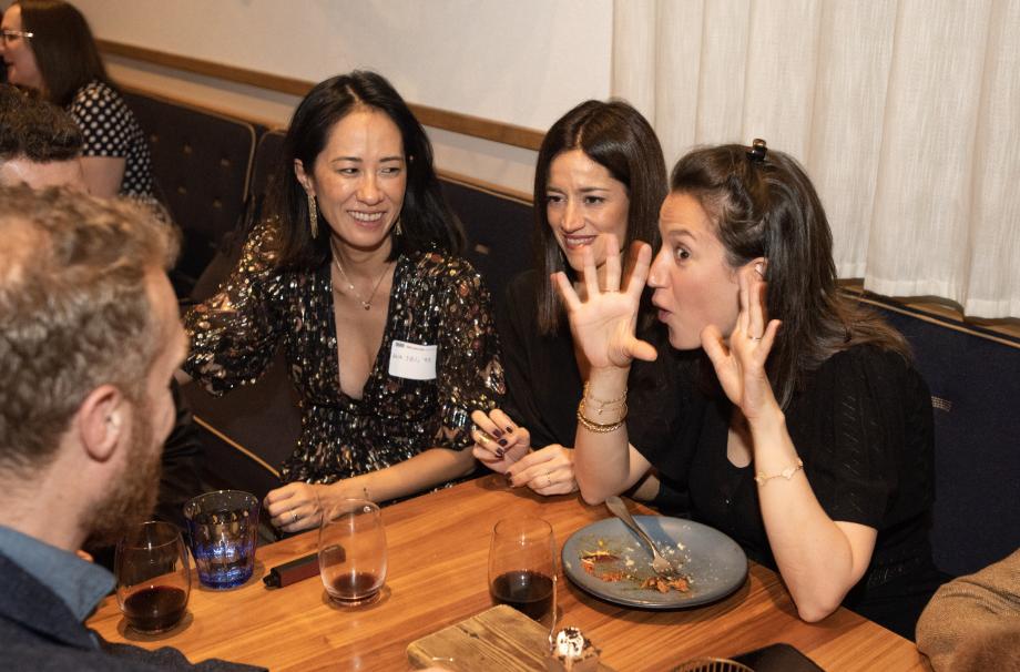 A woman makes a surprised expression while holding her hands up at a man across the table from her. Two other women look on with slight smiles.