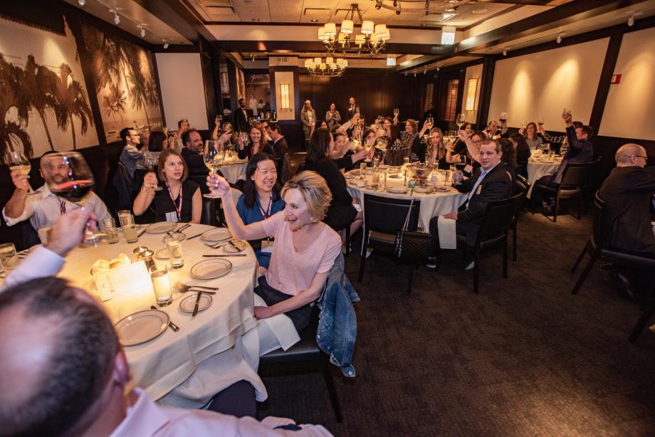 Dozens of people at round tables in a restaurant hold their wine glasses aloft in preparation to cheers one another.