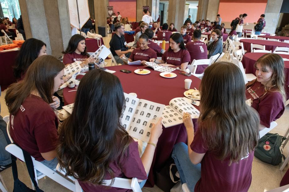 New students in matching maroon shirts sit around large circular tables with books open in front of them.