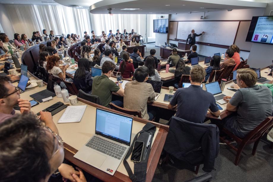Dozens of new students sit in a classroom with laptops and tablets open in front of them while the professor at the front of the room marks a whiteboard.
