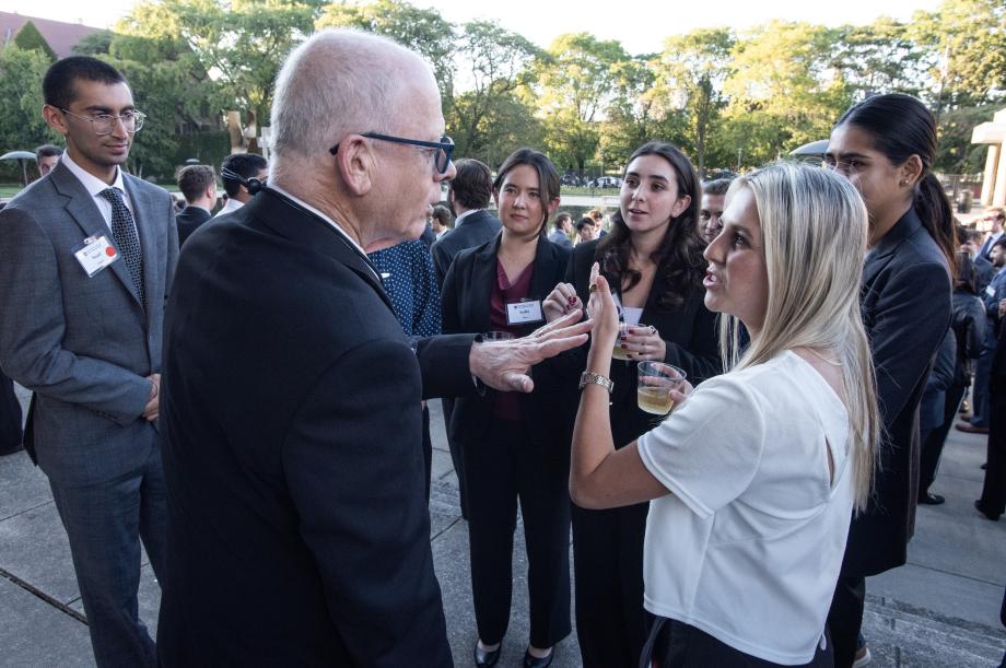 Approximately five new students in professional attire gather in a semi-circle around Saul Levmore, in a suit. One student gestures in conversation with Levmore, who also gestures.