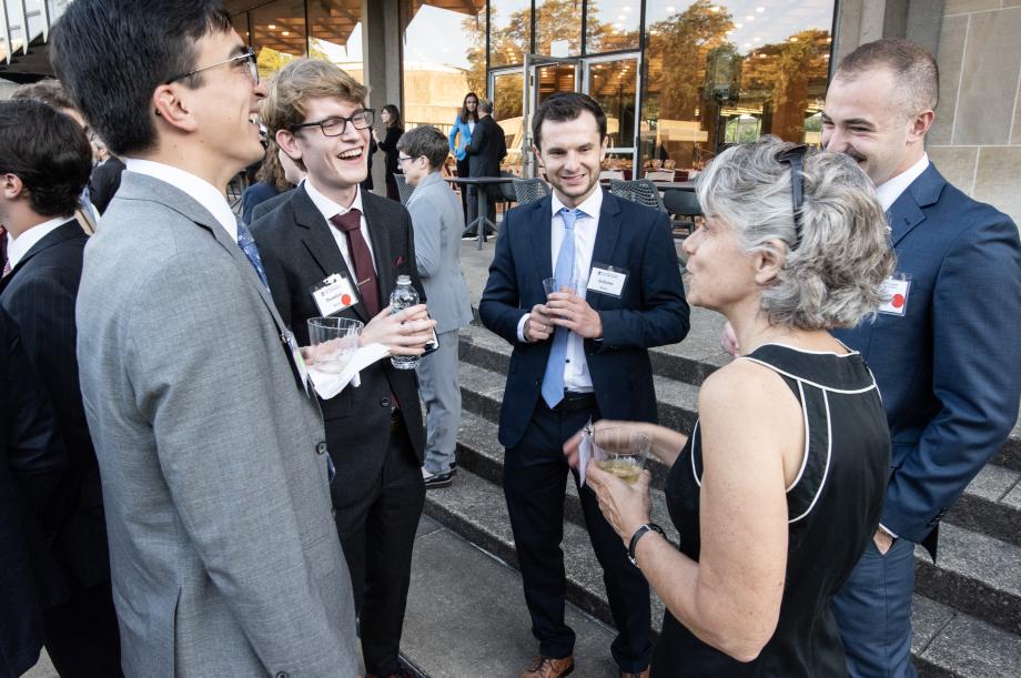 Four new students in suits gather in a semi-circle around Emily Buss, dressed professional. The students laugh as Buss speaks.