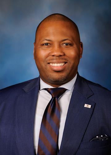A photo of Representative Kam Buckner, the speaker for this event. Representative Buckner is smiling and wearing a blue suit with a red and blue tie.