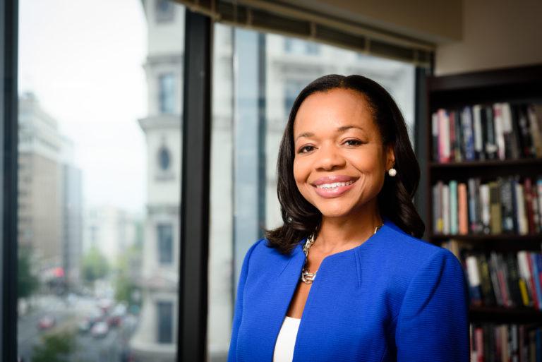 A photo of President Kristen Clarke. She is smiling, and wearing a blue blazer with a white shirt. Behind her there is a city skyline and a bookshelf.