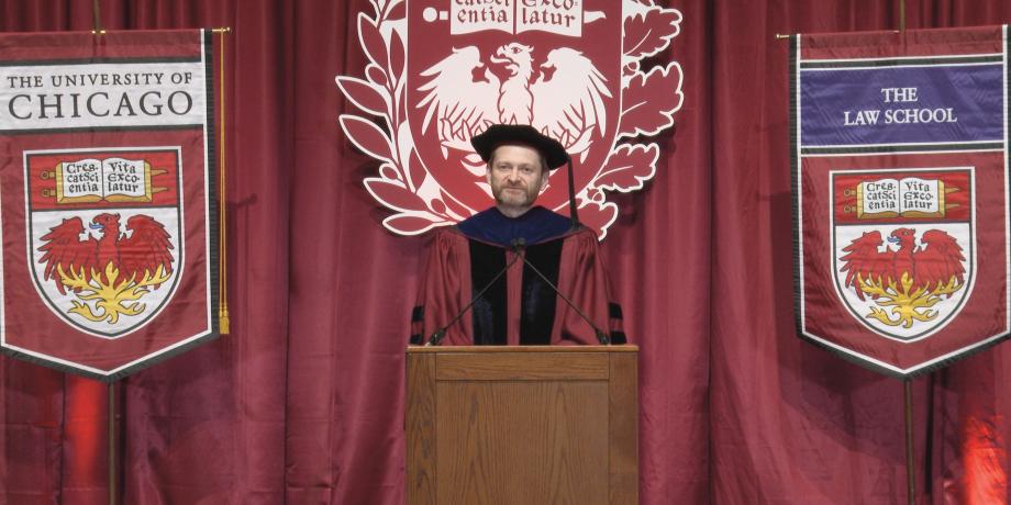 Dean Miles in regalia speaking in front of graduation banners