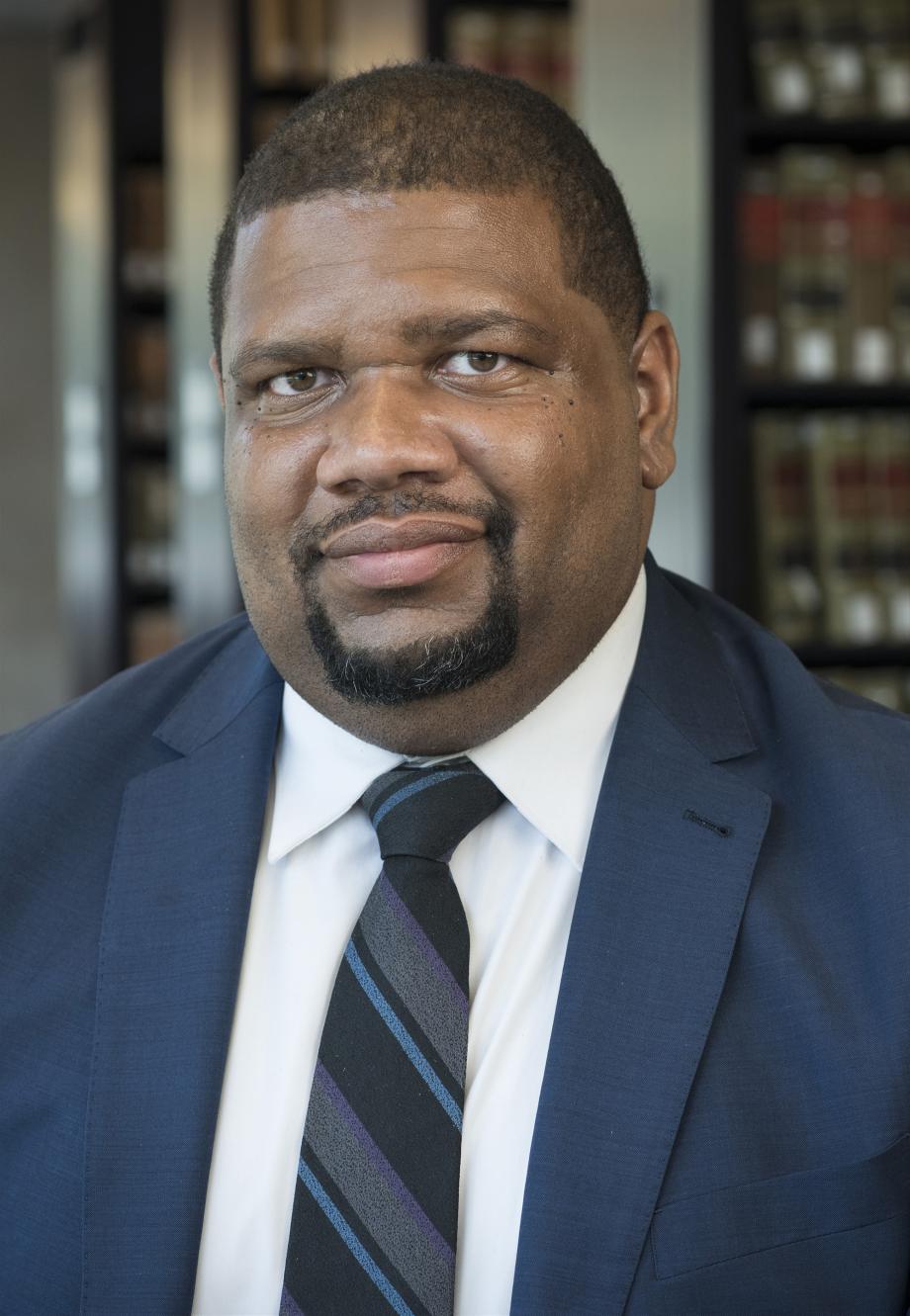 A full color photograph of Professor Adam Davidson, wearing a suit and tie, standing in front of bookshelves.